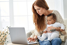 Busy Young Single Parent Caucasian Mother Using Laptop Computer Distance Working Online From Home Office Having Video Call Virtual Meeting With Little Cute African American Infant Baby Girl Daughter.