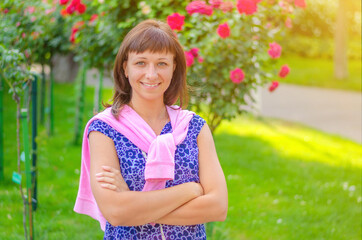 Young beautiful caucasian girl with dark hair and flowered shirt looking at camera, posing, crossing arms and smile in garden with flowers and green trees in sunny summer day with sun flare backlit