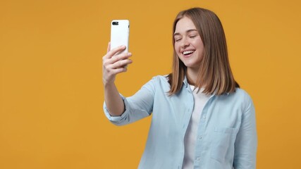 Wall Mural - Cheerful blonde young woman 20s in blue shirt posing isolated on yellow background studio. People lifestyle concept. Talking on mobile cell phone making video call greeting with hand pointing finger