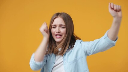 Wall Mural - Cheerful funny excited blonde young woman 20s years old in casual blue shirt posing isolated on yellow color background in studio. People lifestyle concept. Dancing clenching fists waving rising hands