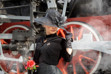 Beautiful girl in a black dress and hat near an old steam locomotive and big iron wheels. Blond beauty. Vintage portrait of the last century, retro journey.