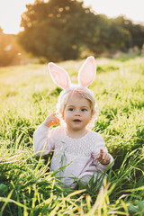 Wall Mural - Happy and cute baby girl wears rabbit ears. Sitting in green grass. The girl has blonde curly hair and a pink dress. Easter concept. Spring time. 