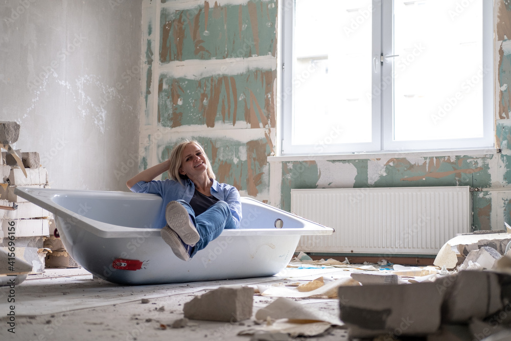 Renovation apartment. Creative story young happy woman sits in bathtub in the middle of the room. Empty walls, repairs house with their own hands. - obrazy, fototapety, plakaty 