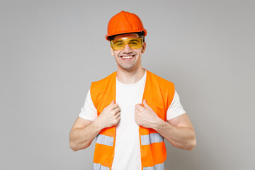 Young employee smiling handyman man in orange protective helmet hardhat look camera touch vest isolated on grey background studio portrait. Instruments for renovation apartment. Repair home concept.