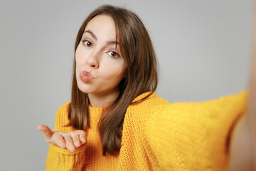 Wall Mural - Close up young smiling happy positive attractive romantic woman 20s in casual knitted yellow sweater do selfie shot on mobile phone blowing air kiss isolated on grey color background studio portrait