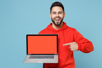 Wall Mural - Young caucasian smiling web designer bearded man 20s in casual red orange hoodie point index finger on laptop pc computer with blank screen workspace area isolated on blue background studio portrait.