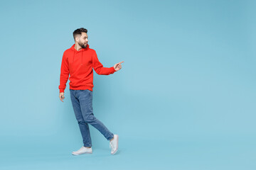 Wall Mural - Full length of young caucasian smiling bearded man 20s ?? casual red orange hoodie pointing on workspace area copy space mock up isolated on blue background studio portrait People lifestyle concept.