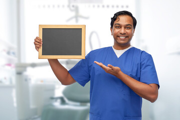 healthcare, dentistry and medicine concept - happy smiling indian male doctor or dentist in blue uniform with chalkboard over dental clinic background