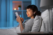 technology, internet and people concept - happy smiling young asian woman with smartphone and wireless earphones lying in bed at home at night
