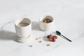 Breakfast, snack still life scene. Cup of coffee, red grapes fruit on white marble table in sunlight. Bowl with muesli, granola and oat cereals and black spoon. Healthy eating, diet concept. Top view.