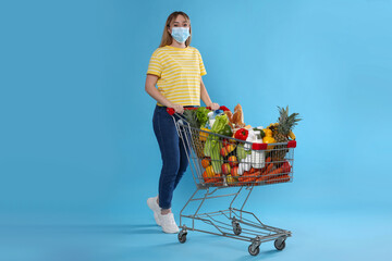 Poster - Young woman in medical mask with shopping cart full of groceries on light blue background