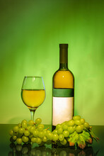 A Green Bottle Of White Wine Next To A Glass Filled With Wine, A Bunch Of Grapes In Front. On A Green Background