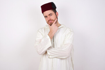 Sticker - Thoughtful smiling young handsome Caucasian man wearing Arab djellaba and Fez hat over white wall keeps hand under chin, looks directly at camera, listens something with interest. Youth concept.