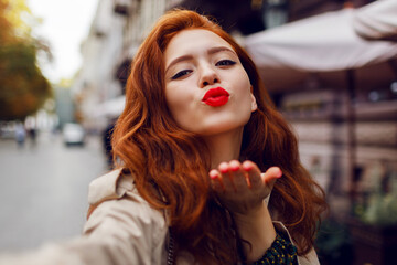 Appealing ginger girl in stylish beige coat walking in cozy European city. Bright make up, red nails, curly hairs.