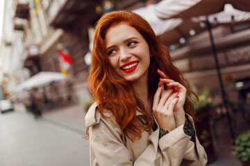 Appealing ginger girl in stylish beige coat walking in cozy European city. Bright make up, red nails, curly hairs.