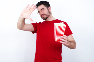 Wall Mural - Portrait of smiling young handsome man in red T-shirt against white background eating popcorn looking at camera and gesturing finger frame. Creativity and photography concept.