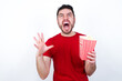 © Jihan - young handsome man in red T-shirt against white background eating popcorn crying and screaming. Human emotions, facial expression concept. Screaming, hate, rage.