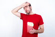 © Jihan - young handsome man in red T-shirt against white background eating corn wiping forehead with hand making phew gesture, expressing relief feels happy that he prevented huge disaster. It was close enough