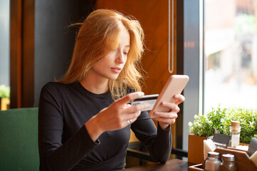 Red haired young woman making card payment through mobile phone to pay bills.