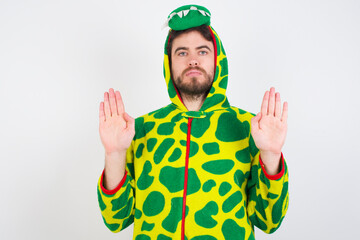 Sticker - Serious young caucasian man wearing a pajama standing against white background pulls palms towards camera, makes stop gesture, asks to control your emotions and not be nervous