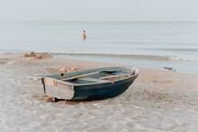 Boat With A Paddle On A Sandy Beach By The Sea. Close-up, At Dawn. A Woman In The Sea And A Seagull In The Background. The Image Is In Pastel Colors. Horizontal Photo
