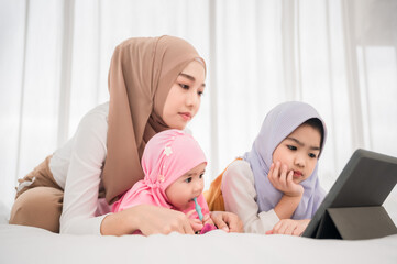 asian muslim mother and daughter in hijab sitting resting on bed and see technology video from table