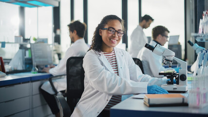 medical science laboratory: beautiful black scientist is using microscope, looking at camera and smi