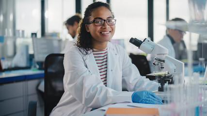 Wall Mural - Medical Science Laboratory: Beautiful Black Scientist is Using Microscope, Looking at Camera and Smiling Charmingly. Young Biotechnology Science Specialist, Using Technologically Advanced Equipment.