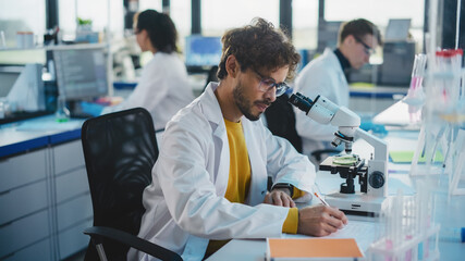 Wall Mural - Medical Science Laboratory: Handsome Latin Scientist is Using Microscope for Sample Analysis and Writes Down the Result, Data. Young Biotechnology Specialist, Using Advanced Equipment.