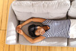 © DimaBerlin - African American young woman wear stripped t-shirt sleeping on couch with arms up at home, closing eyes and taking a break. Black millennial girl resting on sofa, top view. Relax, doze, lazy day.