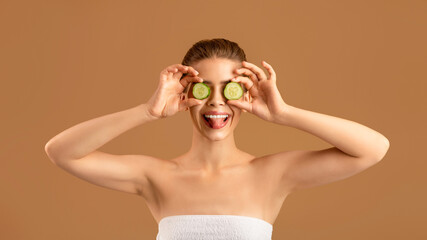 Wall Mural - Natural skincare products. Silly young woman closing eyes with slices of cucumber and showing tongue on brown background