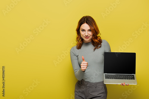 happy woman with curly hair holding laptop with blank screen while showing th...