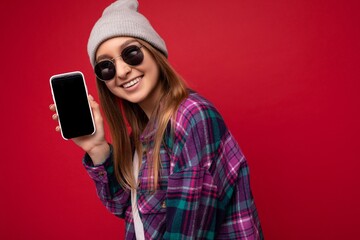 Photo of beautiful smiling young woman good looking wearing casual stylish outfit standing isolated on background with copy space holding smartphone showing phone in hand with empty screen display for