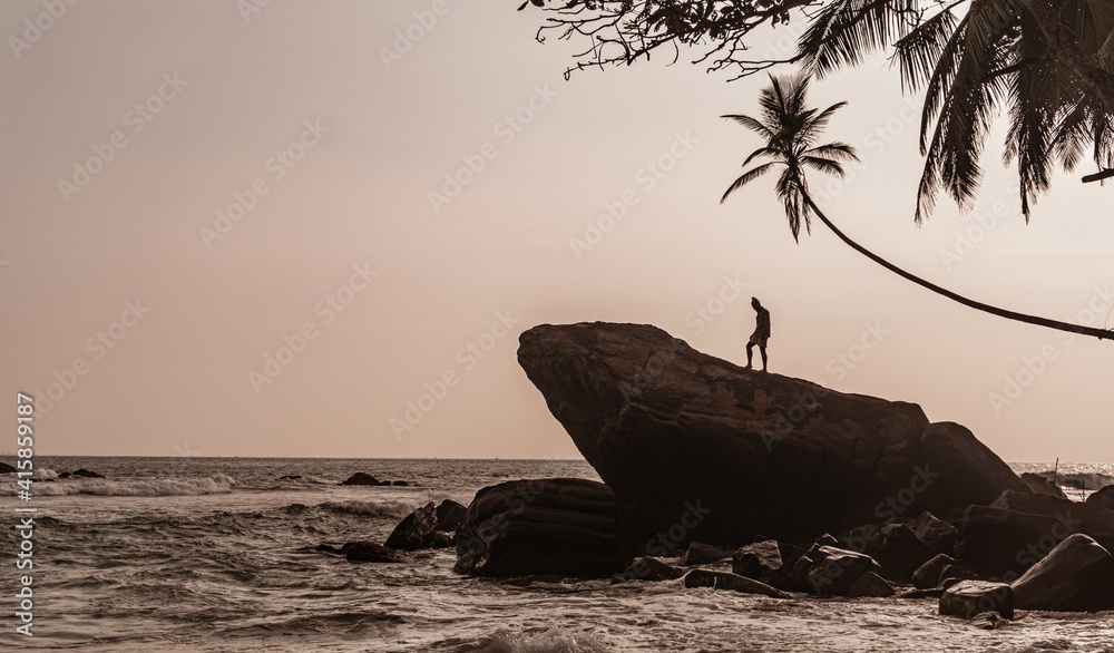 Podróżnik stojący na skale na wybrzeżu oceanu na tle palm i zachodzącego nieba. - obrazy, fototapety, plakaty 