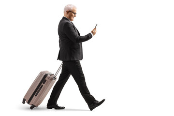 Mature businessman walking and pulling a suitcase while using a smartphone