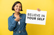 © luismolinero - Young mixed race woman isolated on yellow background holding a placard with text Believe In Your Self and pointing it