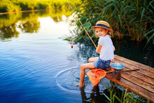 Little Boy In Straw Hat Sitting On The Edge Of A Wooden Dock And Fishing In Lake At Sunset.
