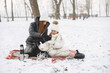 © hetmanstock2 - Family in knitted winter hats on family Christmas vacation. Mother with thermos. People sitting on a blanket.