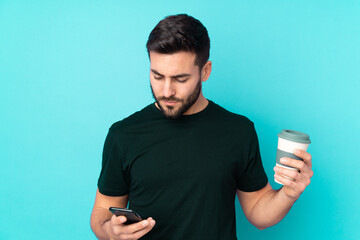 Caucasian handsome man isolated on blue background holding coffee to take away and a mobile