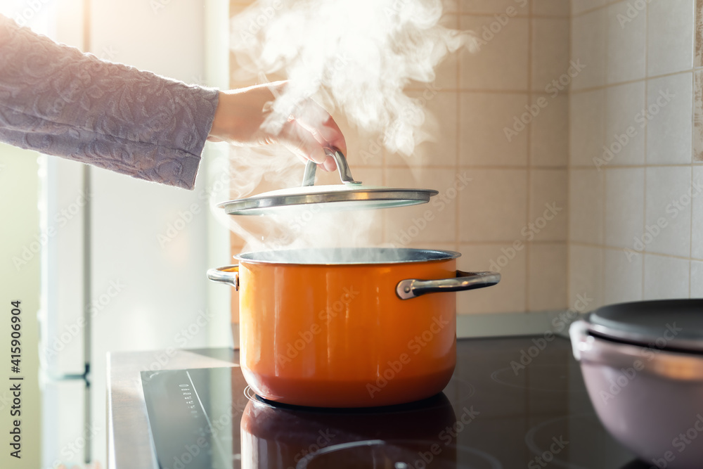 Female hand open lid of enamel steel cooking pan on electric hob with boiling water or soup and scenic vapor steam backlit by warm sunlight at kitchen. Kitchenware utensil and tool at home background - obrazy, fototapety, plakaty 