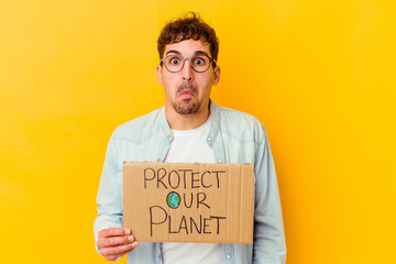 Canvas Print - Young caucasian man holding a protect our planet placard isolated shrugs shoulders and open eyes confused.
