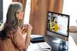 © Kateryna - Online education. Middle aged gray-haired woman studying online via video call uses app and computer, sitting at her work desk, listens to an online lesson, on the screen the teacher near the