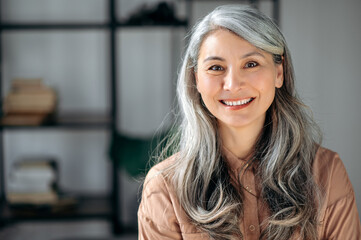 Close-up portrait of a successful mature gray-haired lady, business woman, ceo or business tutor, standing in the office, looking and friendly smiling into the camera