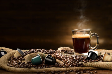 Wall Mural - Cup glass of coffee with smoke and coffee beans and coffee capsules on burlap sack on old wooden background