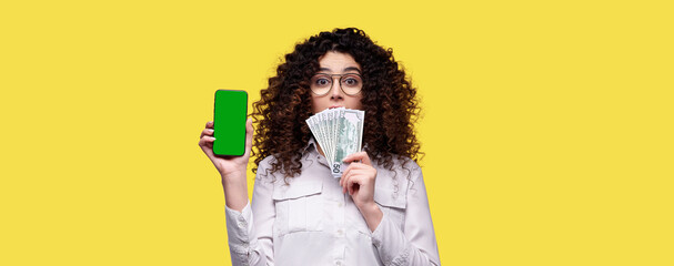 Wall Mural - Surprised woman in glasses holds stack of dollars and smartphone with blank green screen over yellow isolated background. Concept of online casino, bet, games