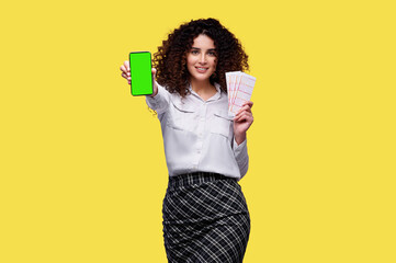 Excited young curly girl holding lottery tickets and smartphone with blank green screen isolated over yellow background