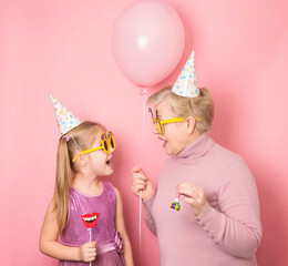 Old granny with her granddaughter in funny eyeglasses and birthday hats having fun together on pink background. Happy birthday concept.