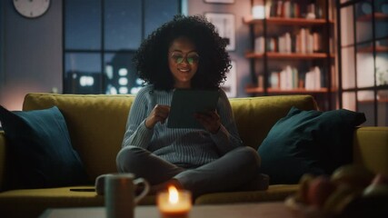 Aufkleber - Beautiful Authentic Latina Female Sitting on Couch Sofa in a Cozy Dark Living Room in the Evening and Using Tablet Computer at Home. She's Browsing the Internet and Checking Videos on Social Networks.