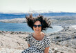 Smiling Caucasian woman in a polka dots flowy dress and sunglasses with hair blown up by the wind