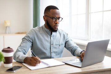 Wall Mural - African Business Guy Working On Laptop Taking Notes At Workplace